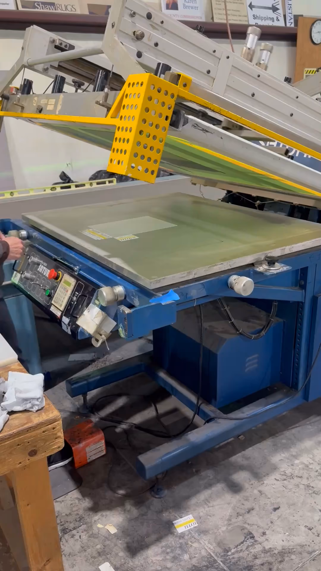 Industrial screen printing machine with yellow platen lifting open on a warehouse floor.