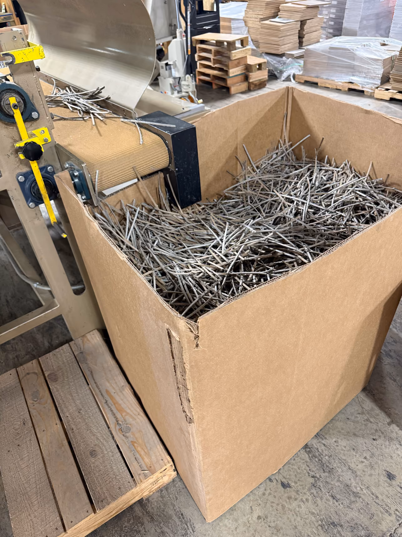 Cardboard box filled with shredded paper strips next to industrial conveyor belt in recycling facility.
