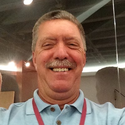 Smiling middle-aged man with a mustache wearing a light blue polo and red lanyard indoors.