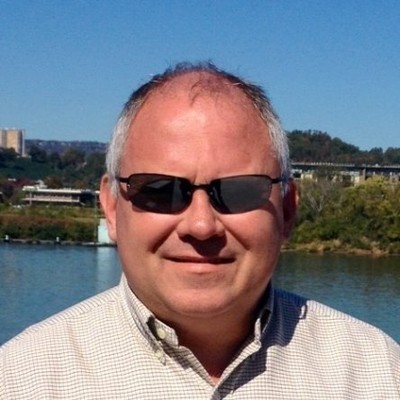 Smiling middle-aged man wearing sunglasses and a checkered shirt near a waterfront on a sunny day.