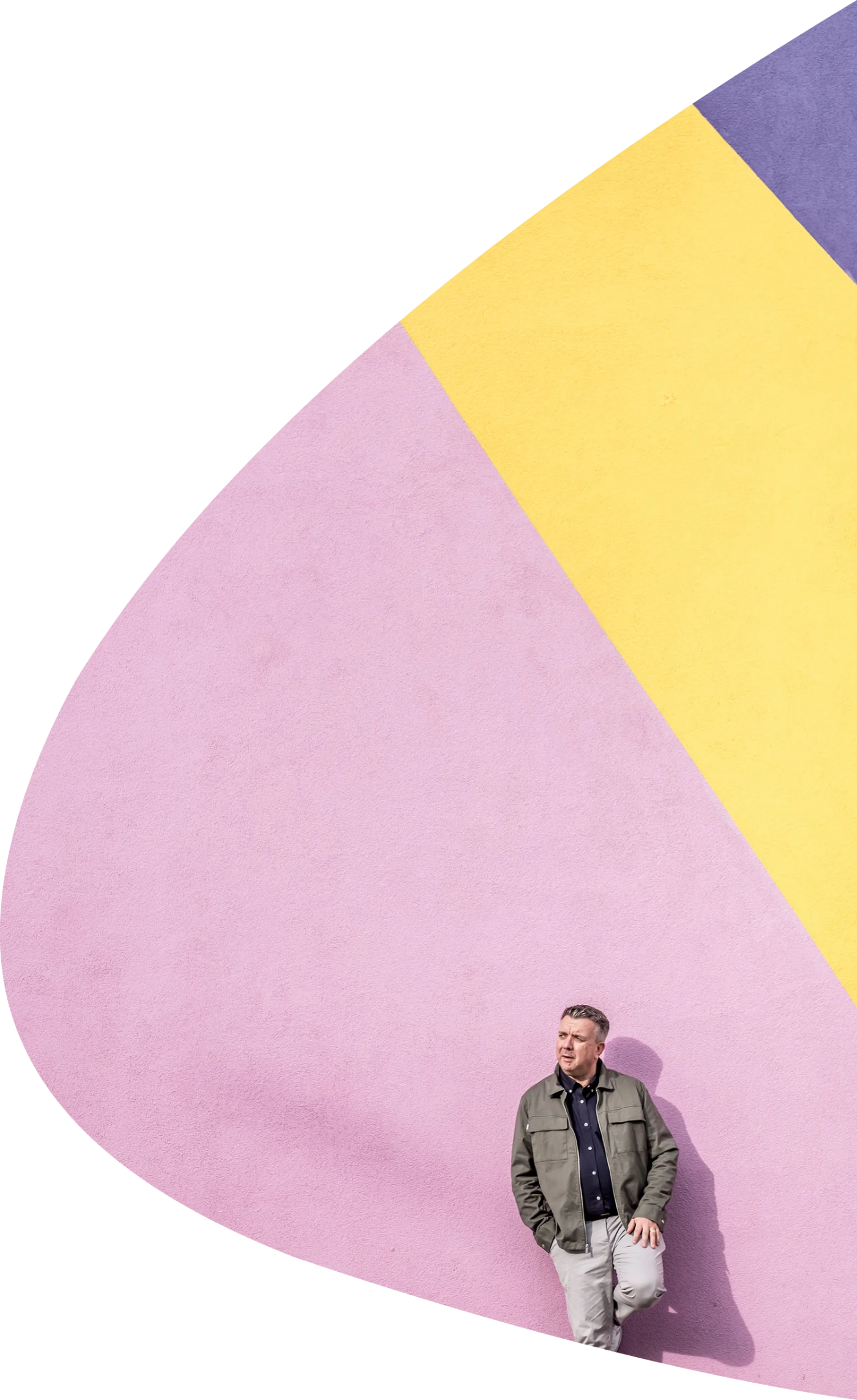 Photograph of Duncan leaning against a bright pink, yellow and purple wall from far away.