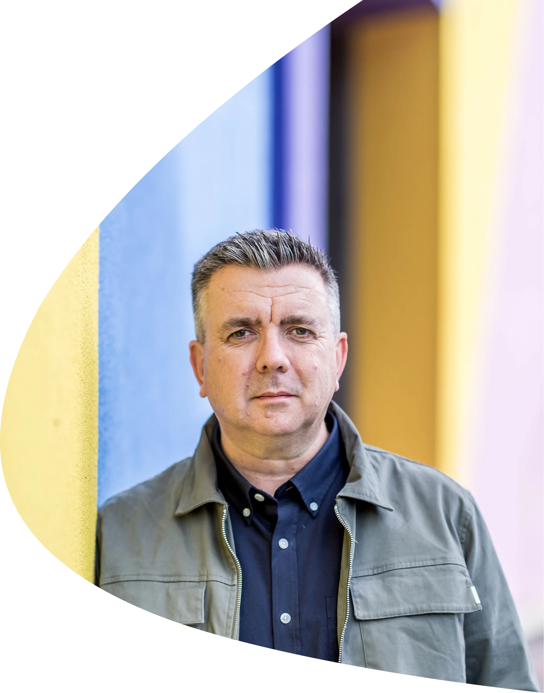 Man with short grey hair wearing a dark shirt and green jacket standing against a colourful wall.