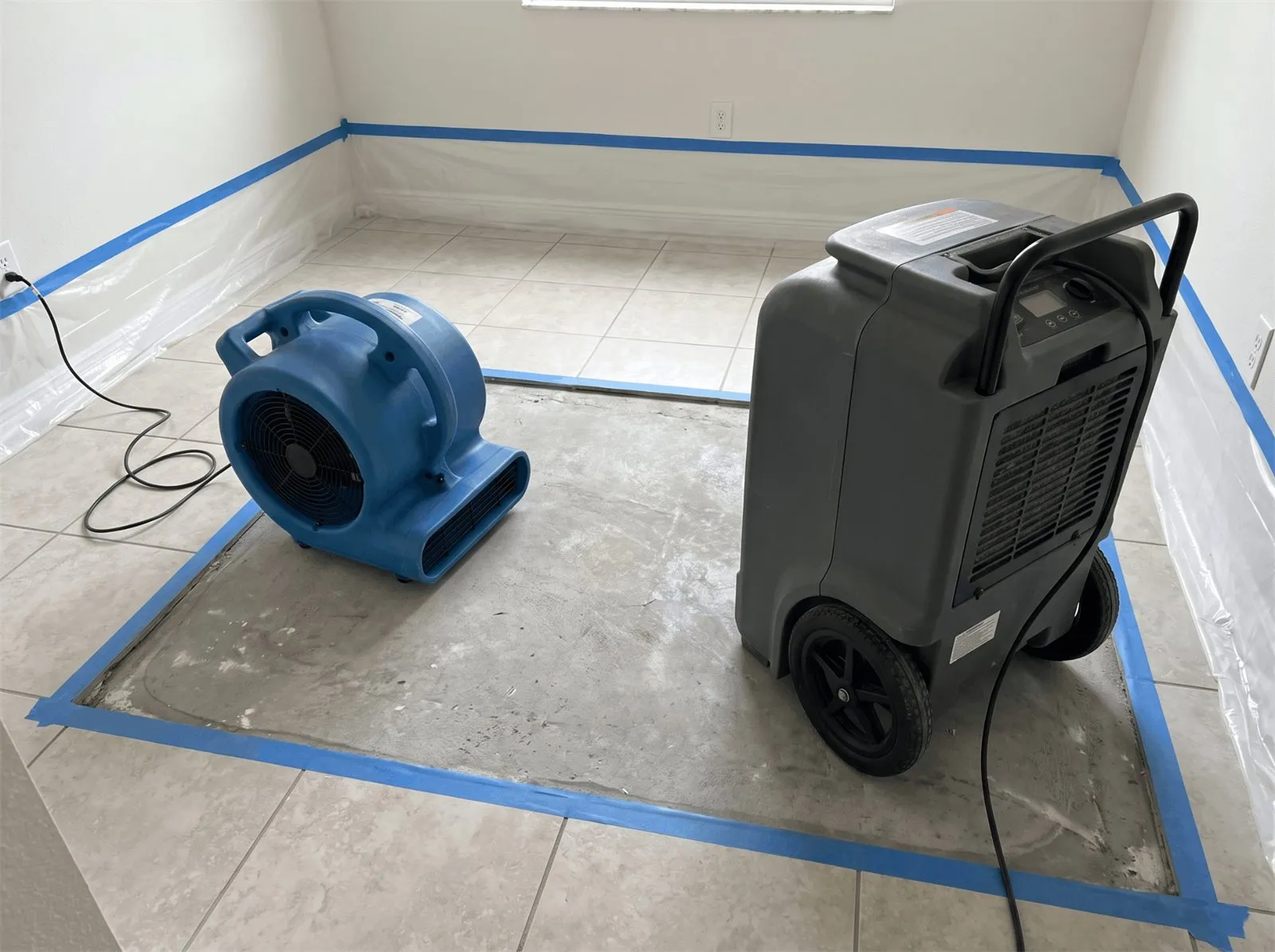 Air mover and dehumidifier drying a taped-off section of tile floor inside plastic containment