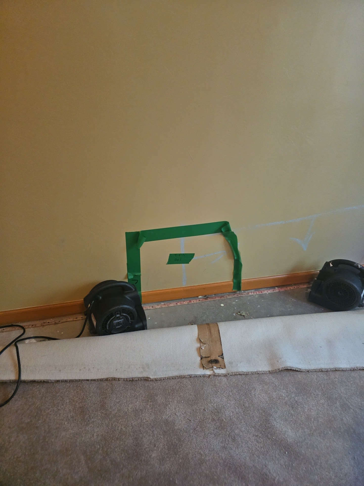 Carpet pulled back from a wall, with two air movers drying the baseboard and a taped-off square marking moisture readings.