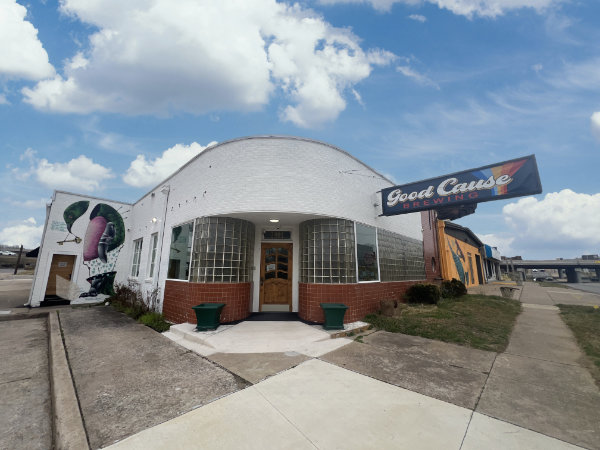 White brick building with a rounded front entrance, a sign reading 'Good Cause Brewing,' and a mural on the side wall under a blue sky with clouds.