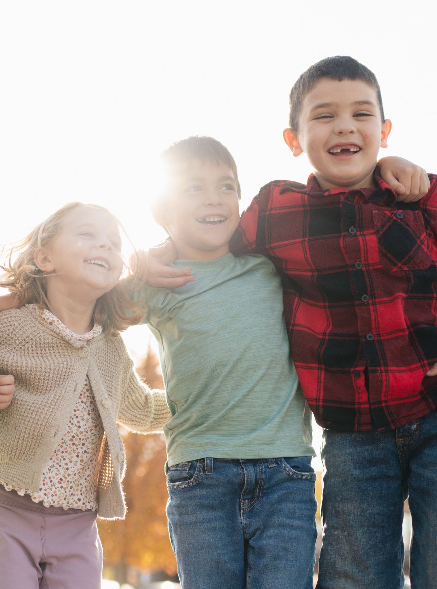 Three children smiling and hugging outdoors with sunlight behind them.