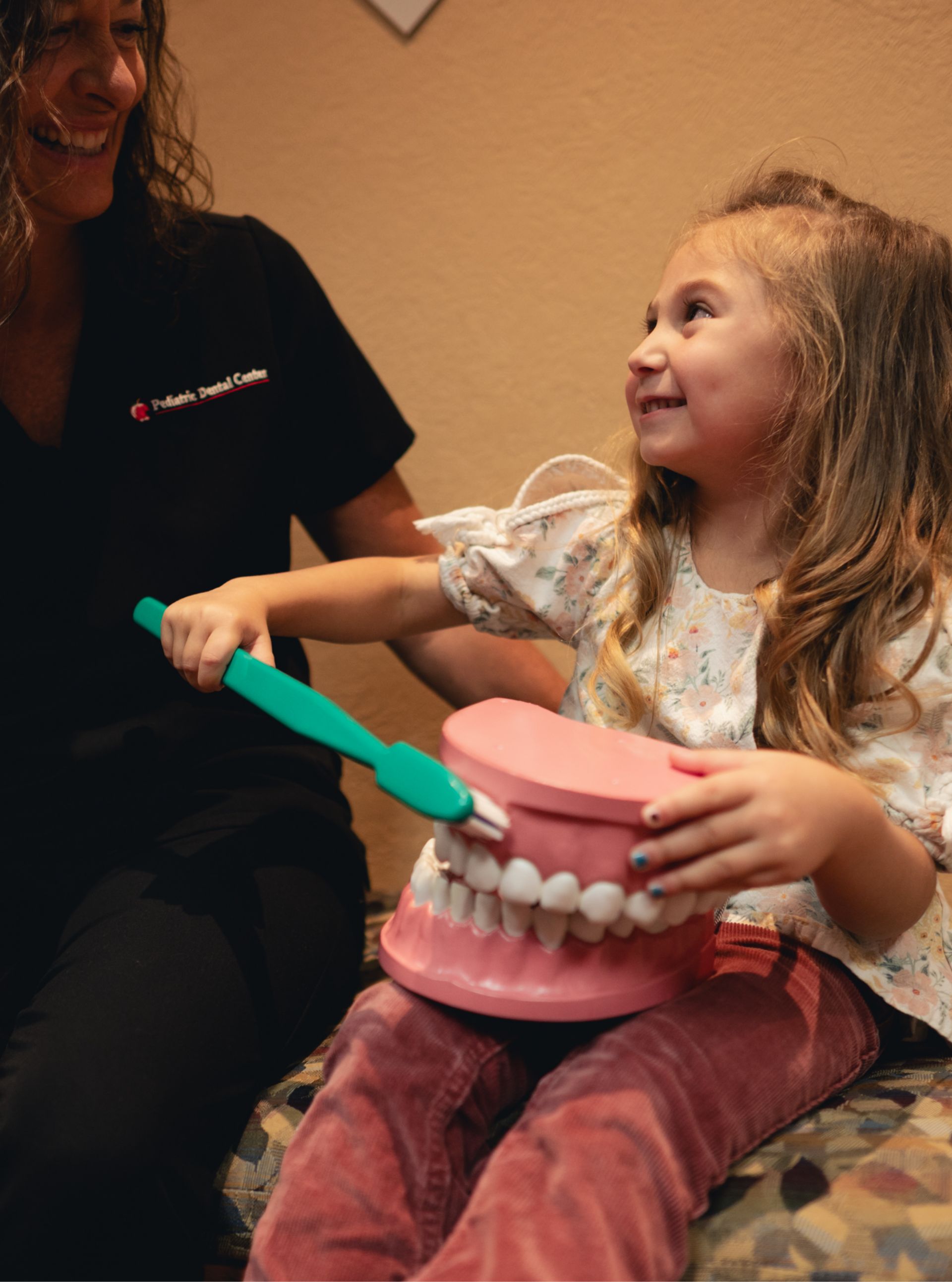 A young girl smiling as she holds a large model of teeth and brushes them with a big toothbrush, sitting next to a woman in a Pediatric Dental Center uniform.