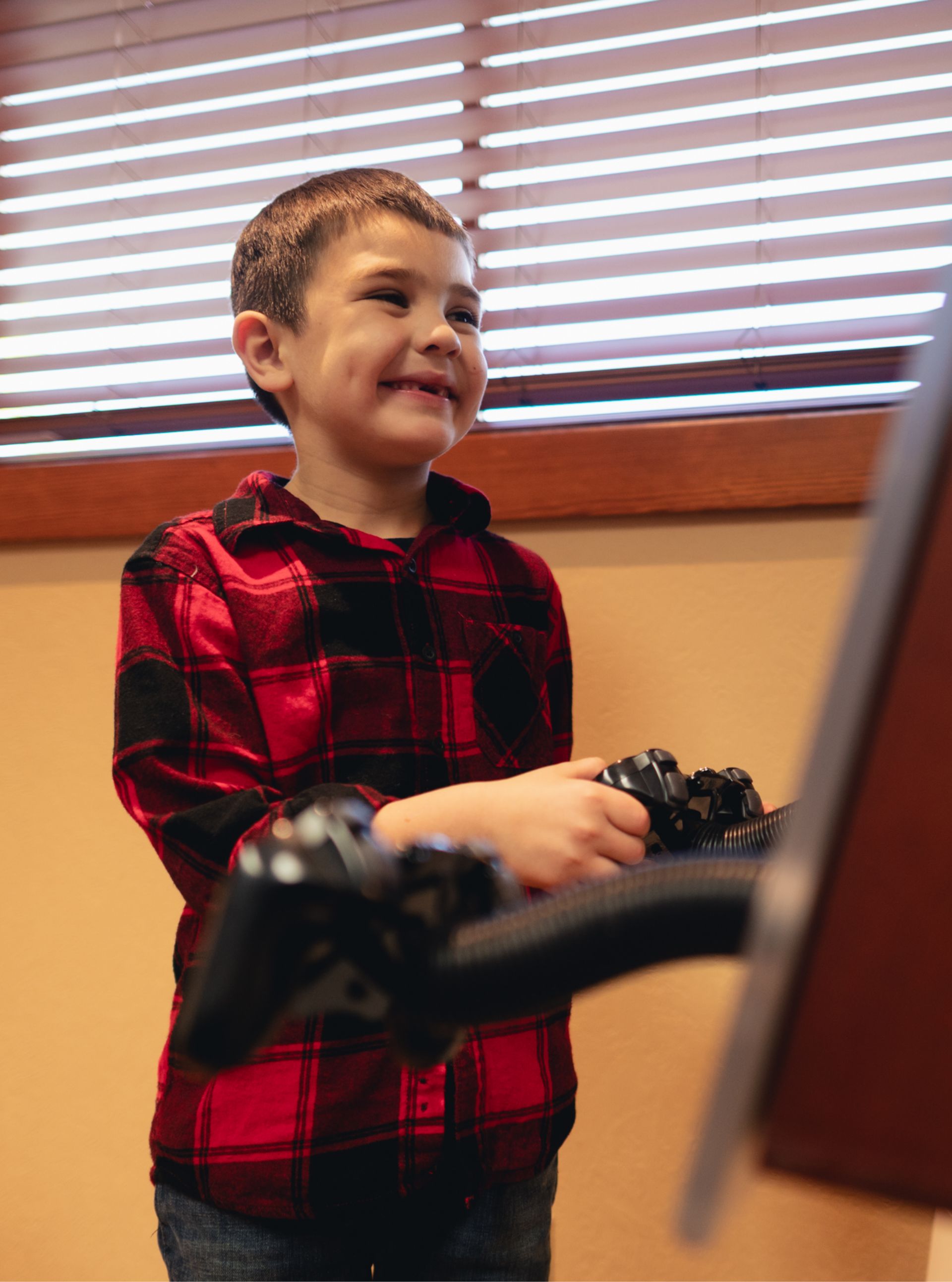 Smiling boy in a red and black plaid shirt playing video games with a controller in hand.