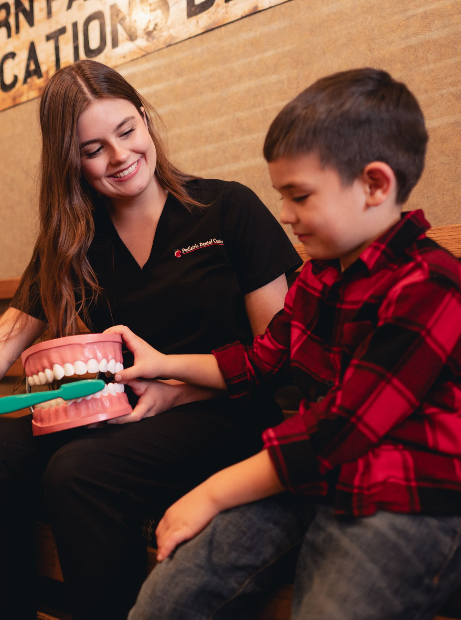 Smiling dental professional teaching a young boy how to brush teeth using a large dental model and a green toothbrush.