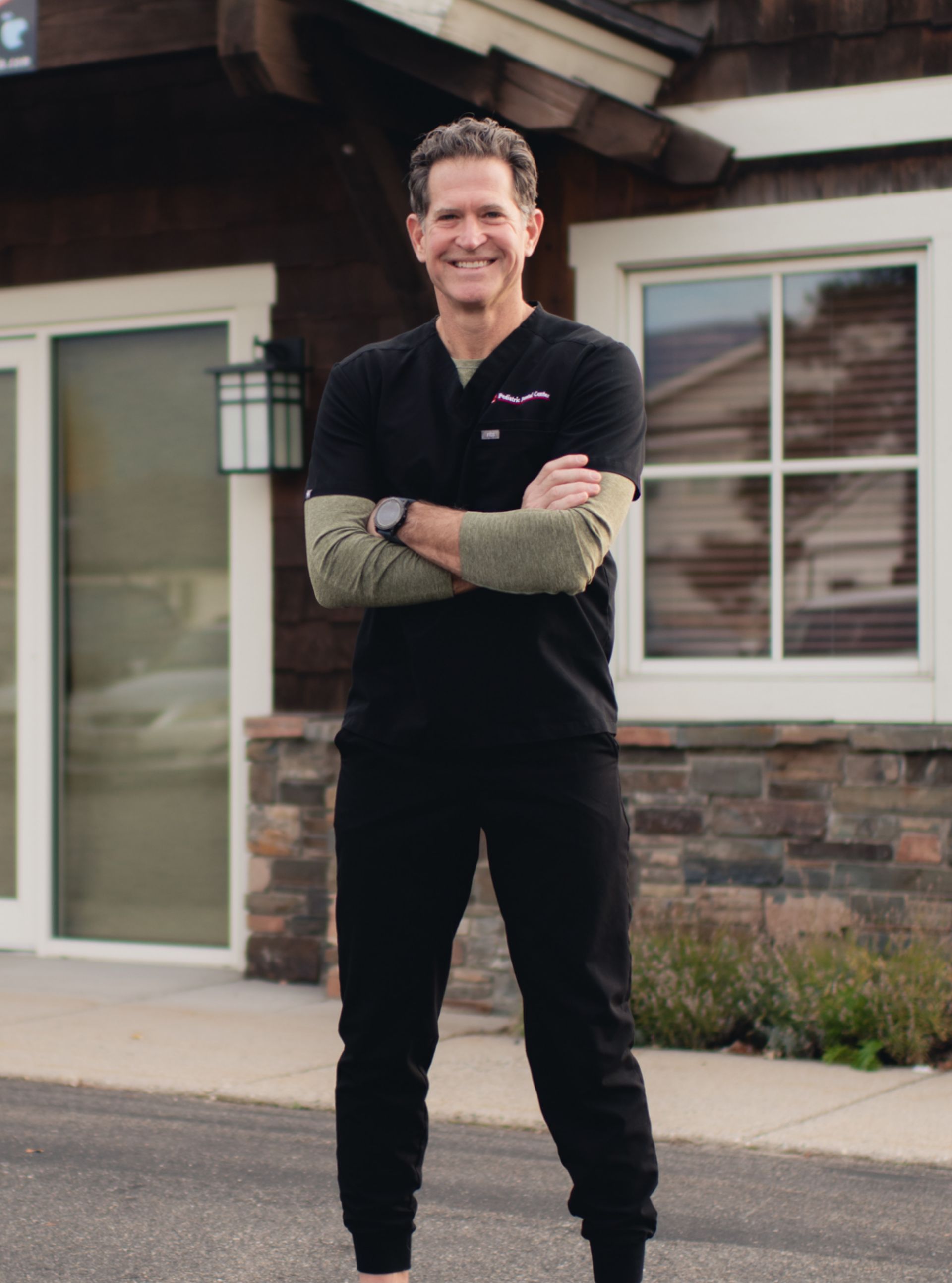 Smiling man in black scrubs and gray long-sleeve shirt standing with arms crossed outside a building with stone walls and windows.