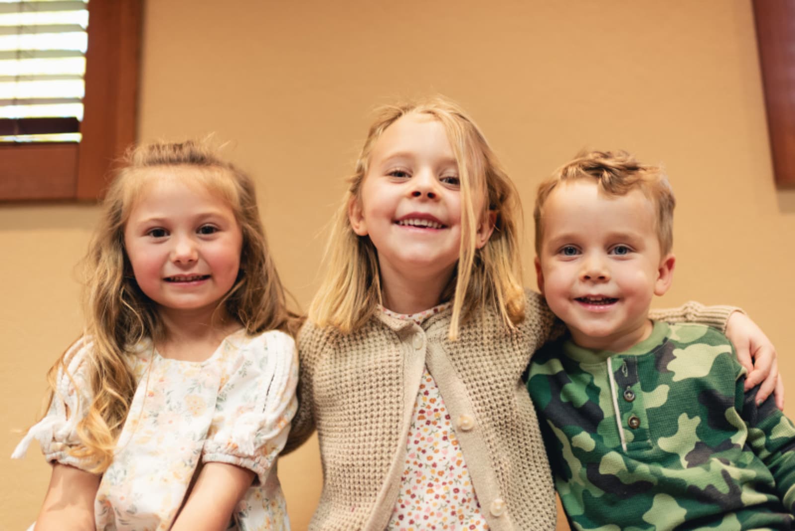 Three smiling children standing together in a dental office setting with a warm beige wall background.
