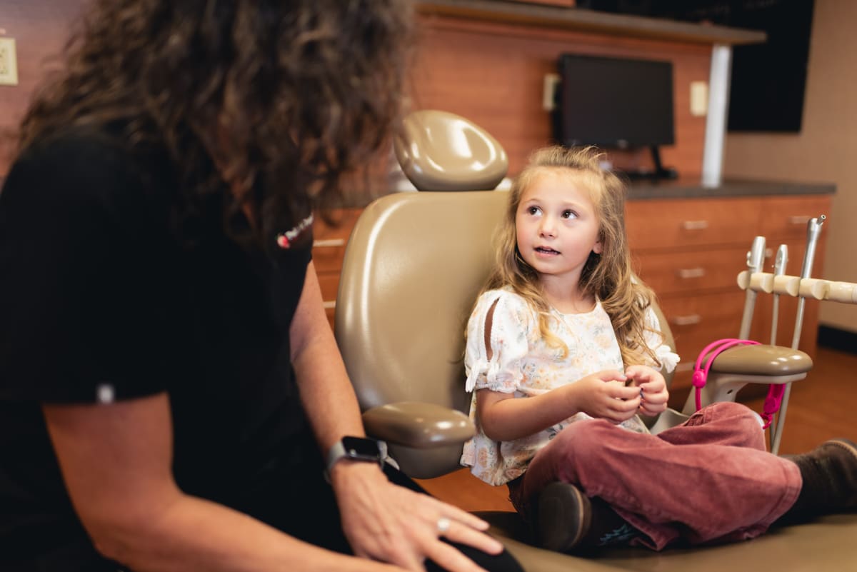A dental professional shows a young child a model or educational tool during a consultation in a warm, wood-toned office sett