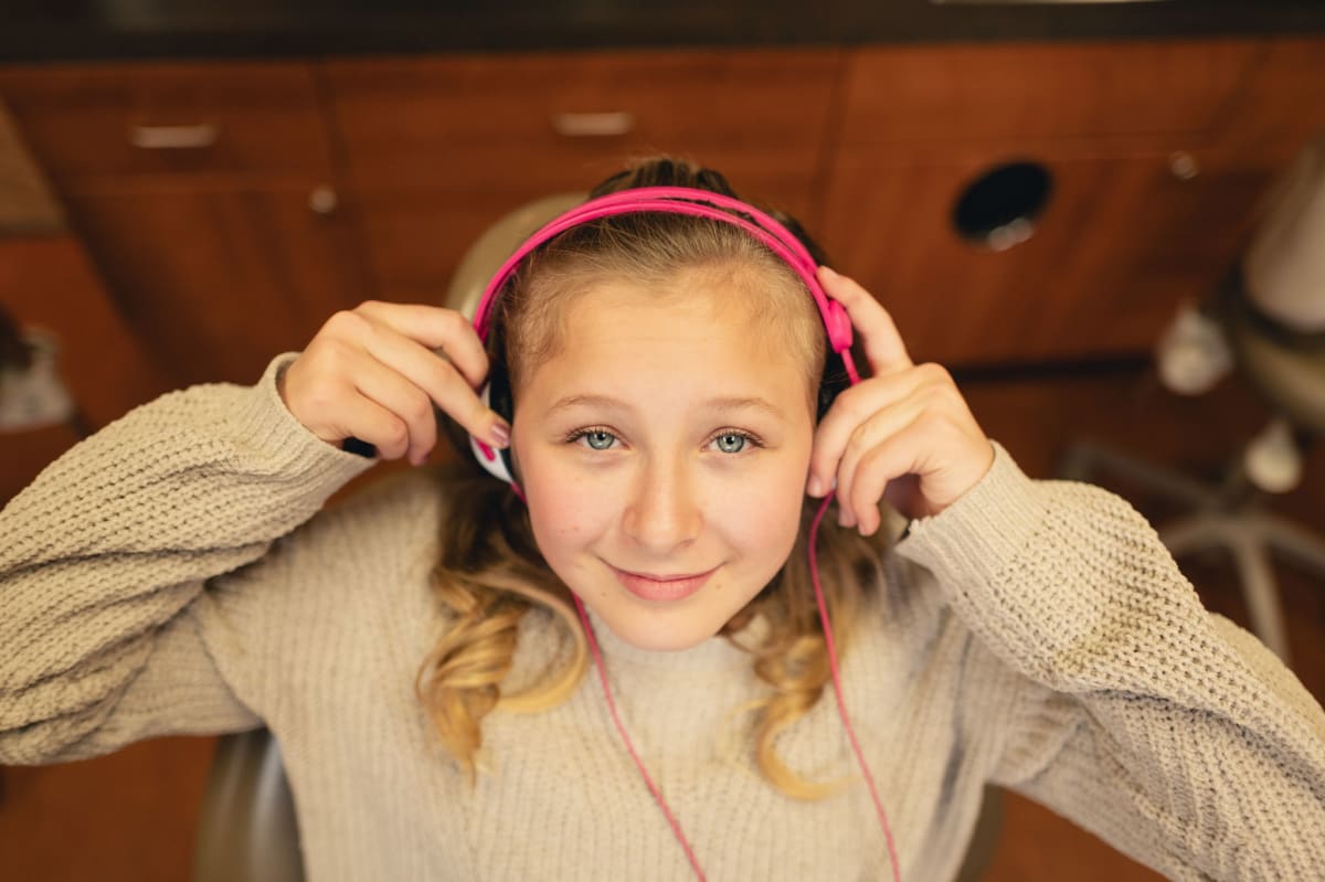 Young girl wearing a pink headband and gray sweater smiling at the camera indoors.
