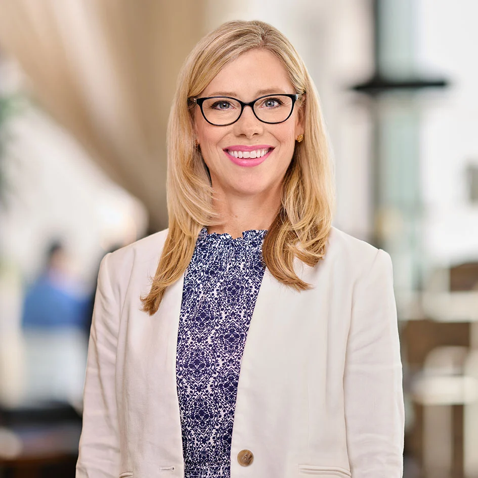 Smiling blonde woman with glasses wearing a white blazer and patterned blouse in a blurred office setting.