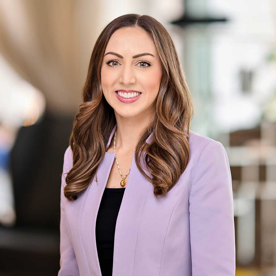 Smiling woman with wavy brown hair wearing a lavender blazer and a gold pendant necklace.
