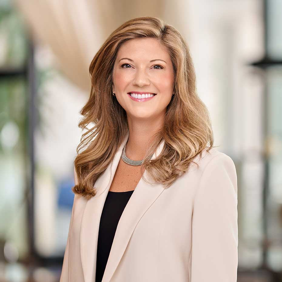 Smiling woman with wavy blonde hair wearing light blazer and silver necklace against blurred background.