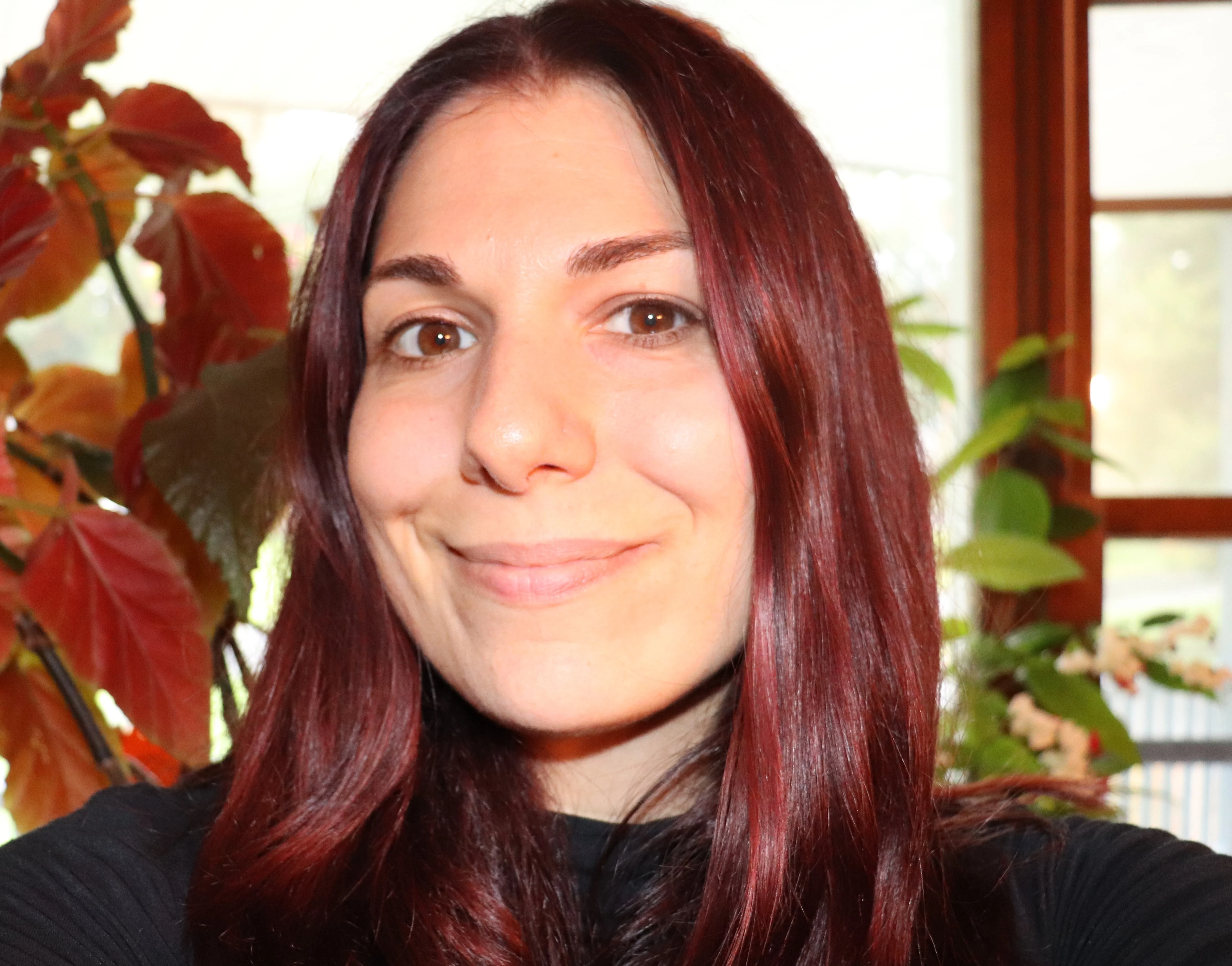 Close-up portrait of a smiling woman with long red hair indoors with plants in the background.