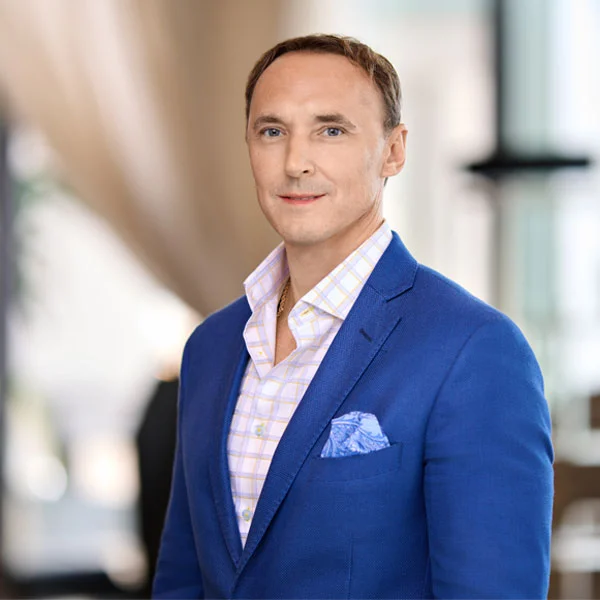 Man wearing a blue blazer and light checkered shirt poses in a softly lit indoor setting.