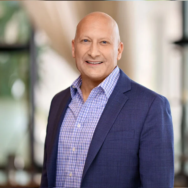 Smiling bald man wearing a blue checkered shirt and navy blazer against a blurred background.