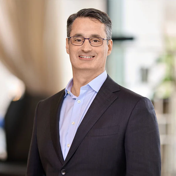 Man with short dark hair wearing glasses, a light blue shirt, and a dark blazer smiling indoors.