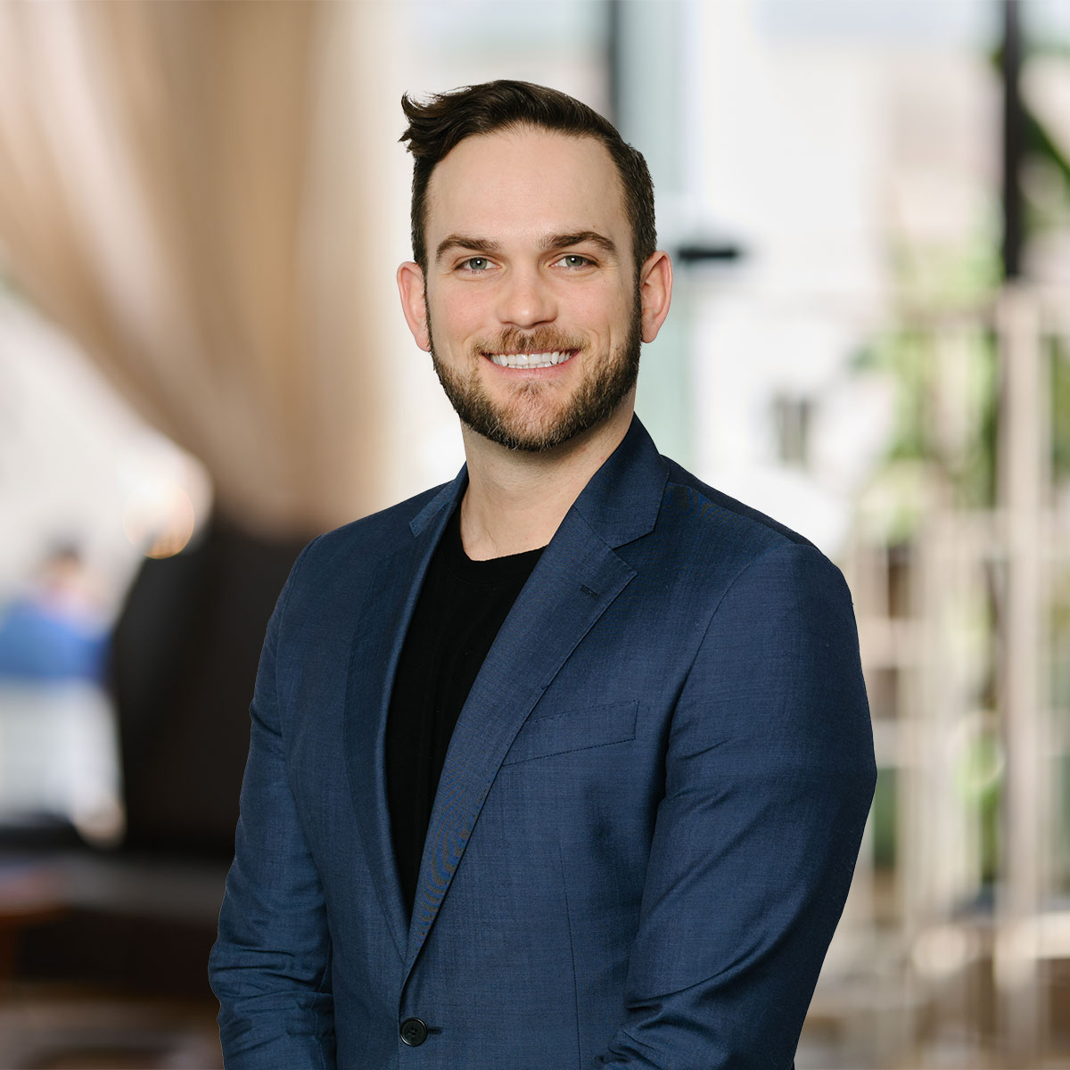 Smiling man with beard wearing a blue blazer and black shirt in a softly blurred indoor setting.