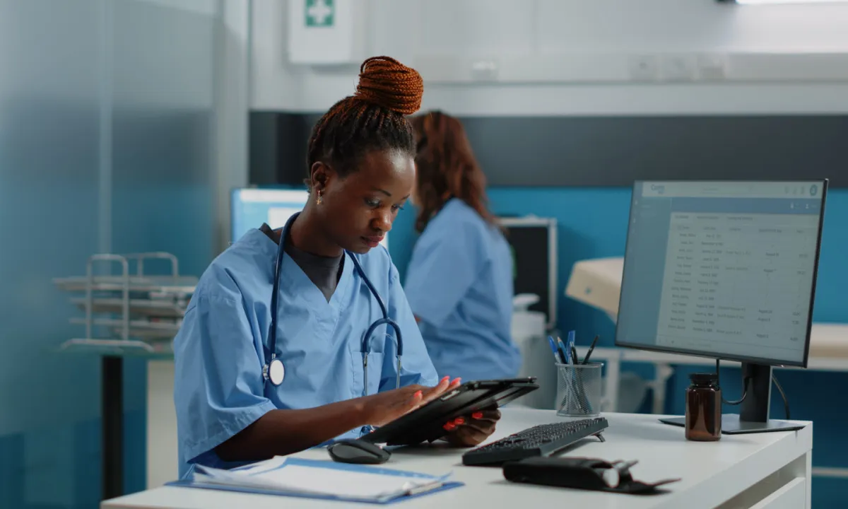 A nurse in blue scrubs uses a tablet at a desk with a computer monitor and medical tools nearby.