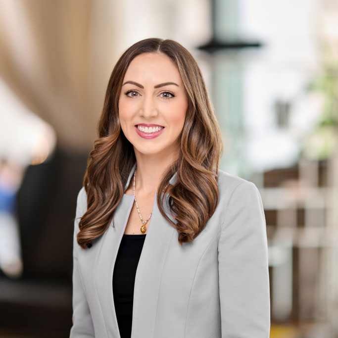 Smiling woman with long wavy hair wearing a gray blazer and black top in a bright indoor setting.