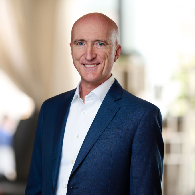 Smiling bald man in a blue suit jacket and white shirt against a blurred background.