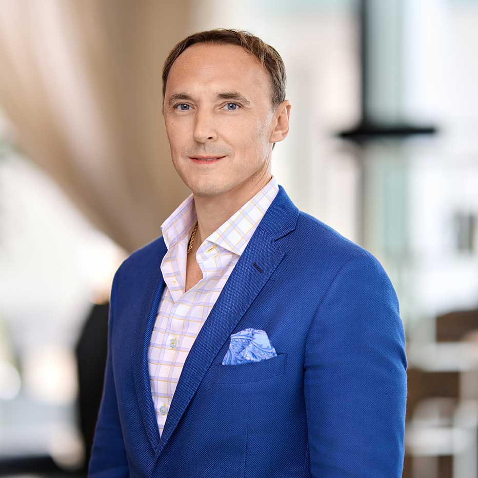 Man in blue suit jacket and checkered shirt standing indoors with a neutral expression.