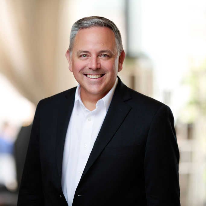 Smiling man with gray hair wearing a black suit jacket and white shirt in a softly lit indoor setting.