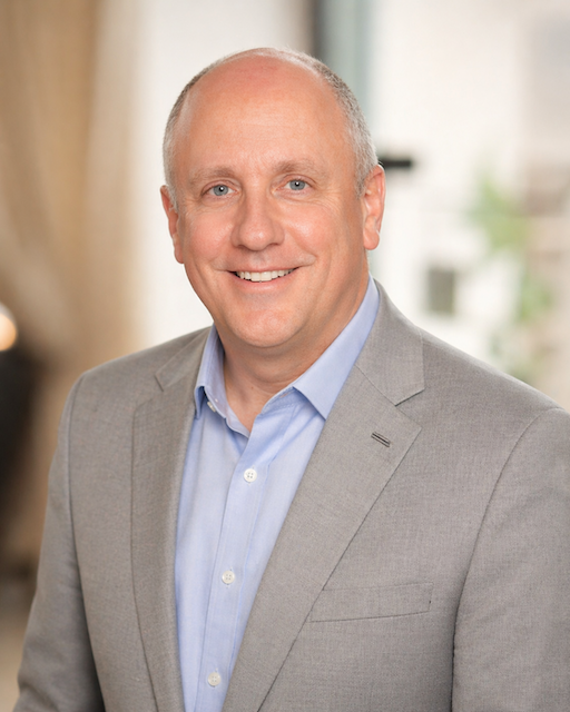 Smiling bald man wearing a light gray blazer and blue shirt in a softly lit indoor setting.