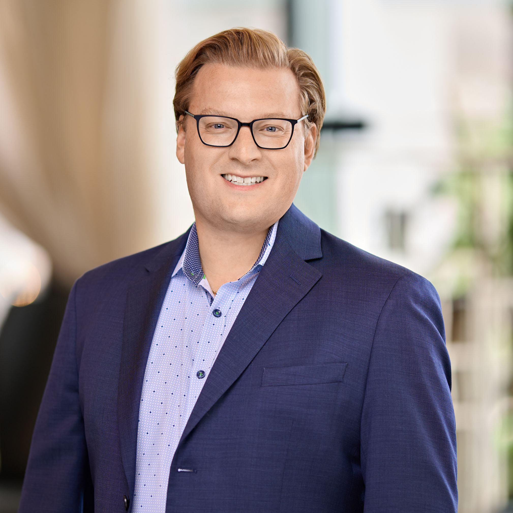 Smiling man with blond hair and glasses wearing a blue suit jacket and light blue shirt.