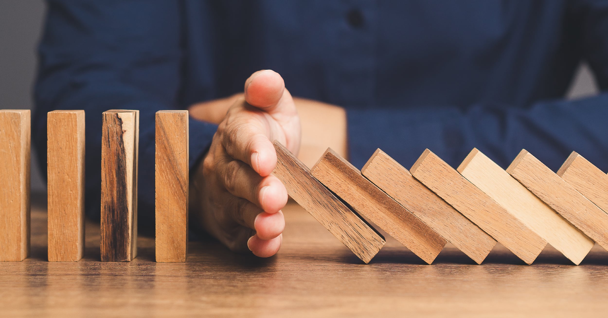 Hand stopping a row of falling wooden domino pieces on a wooden table.