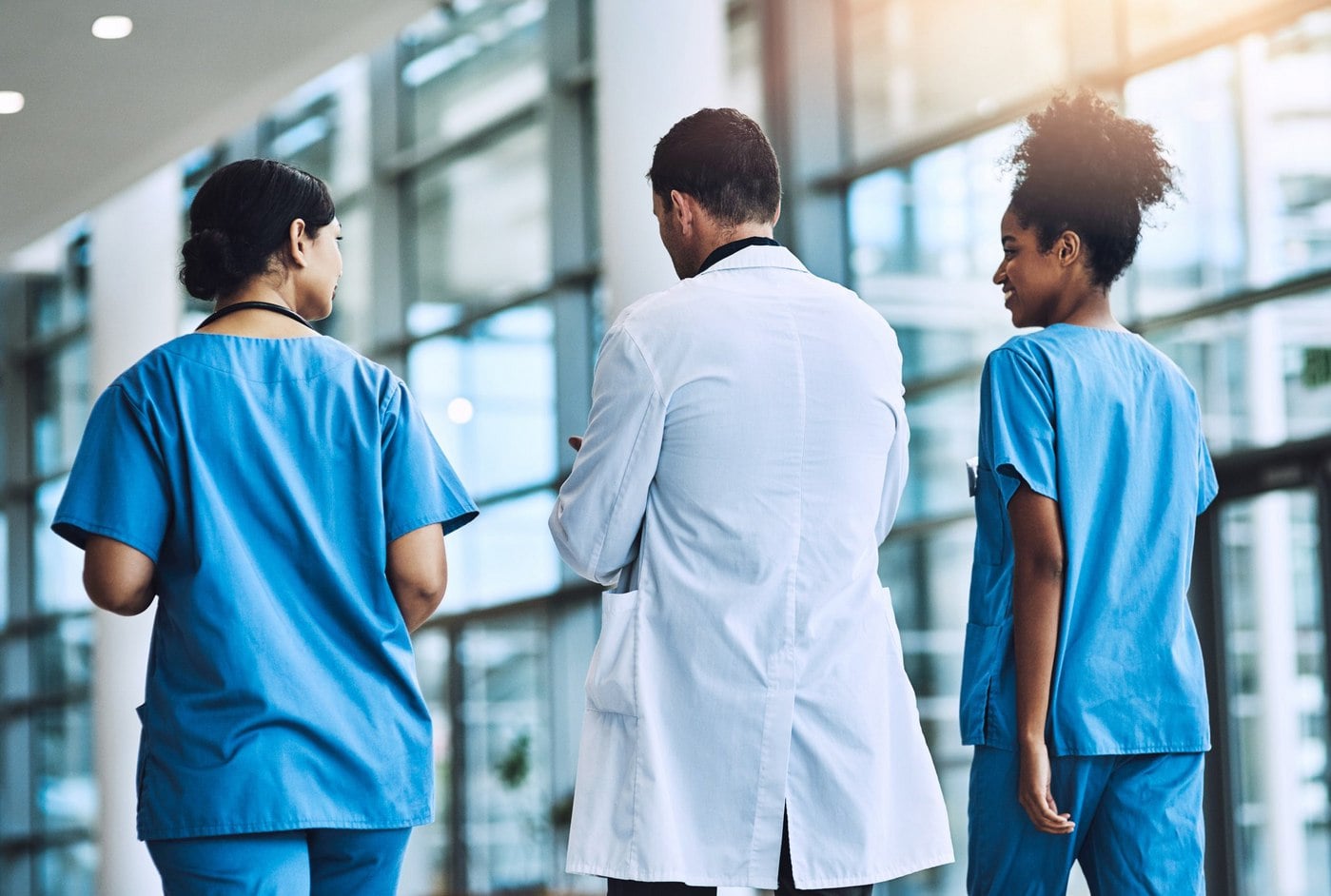 Two nurses in blue scrubs and a doctor in a white coat walking and talking in a hospital corridor.