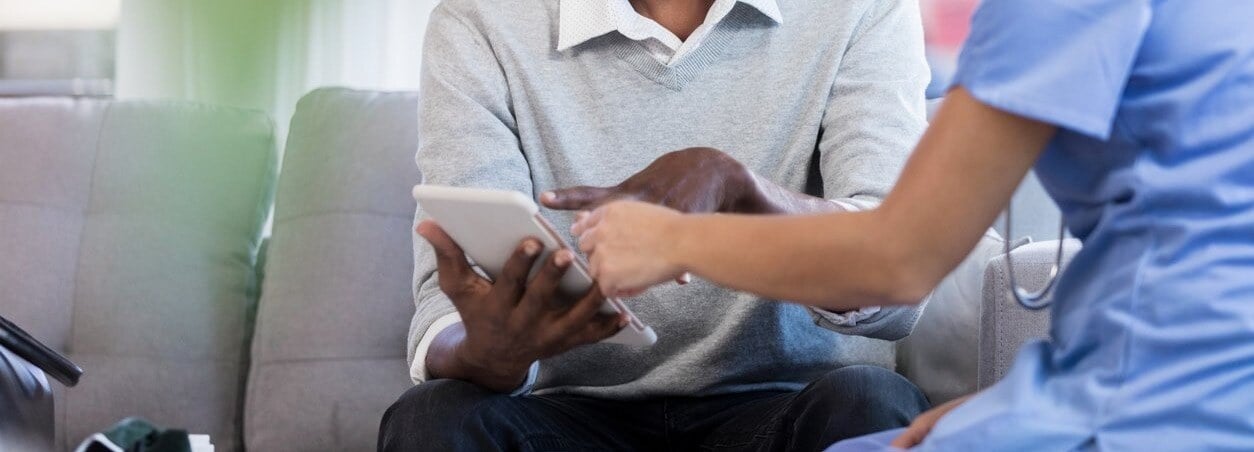 Healthcare worker in scrubs showing information on a tablet to a seated man in casual clothes.
