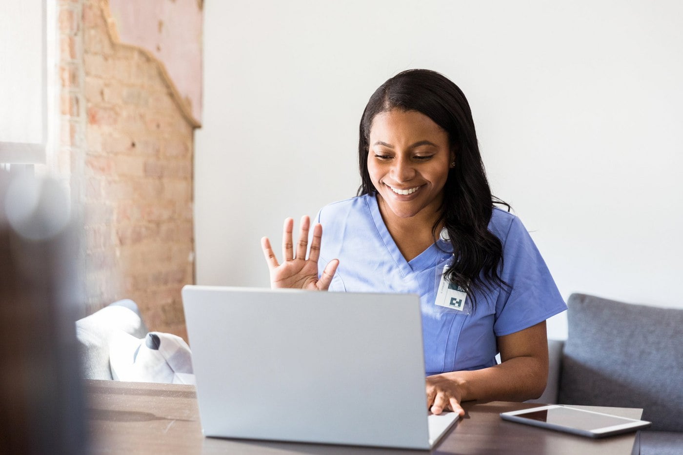 Smiling nurse in scrubs waving during a video call on a laptop at a wooden table.