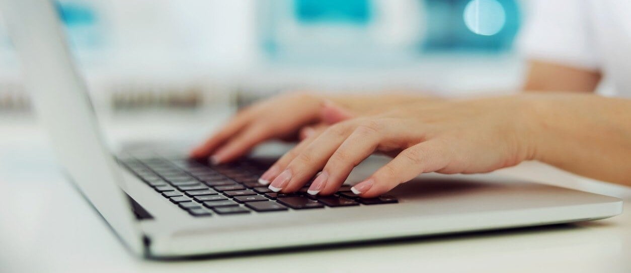 Hands with French manicured nails typing on a laptop keyboard in a bright workspace.
