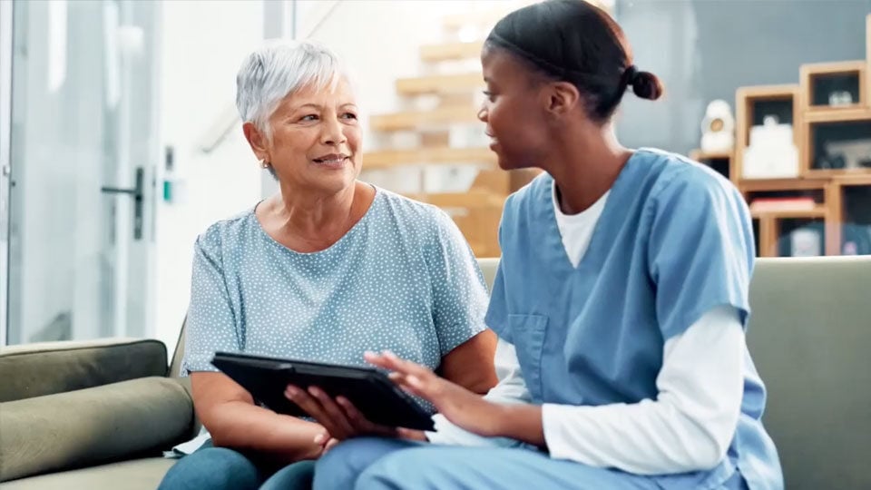 Nurse in blue scrubs talking to an elderly woman holding a tablet on a couch.