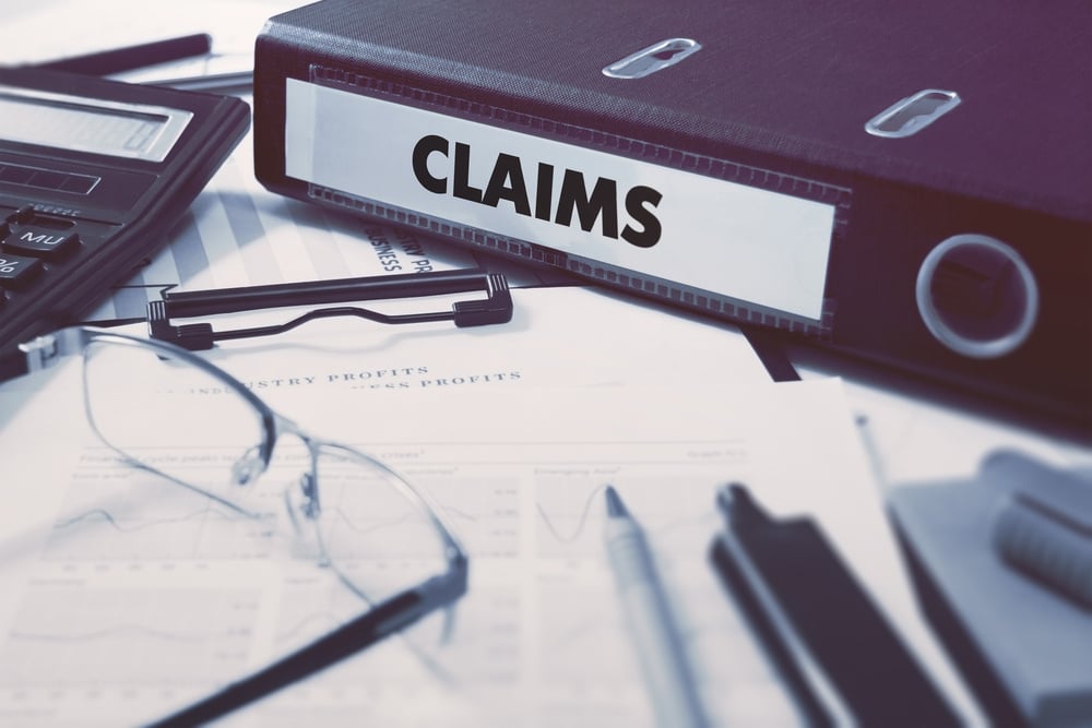 Black ring binder labeled CLAIMS on desk with calculator, glasses, pen, and printed financial documents.
