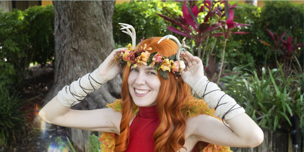 Smiling woman with long red hair wearing a floral crown with small antlers outdoors near greenery.