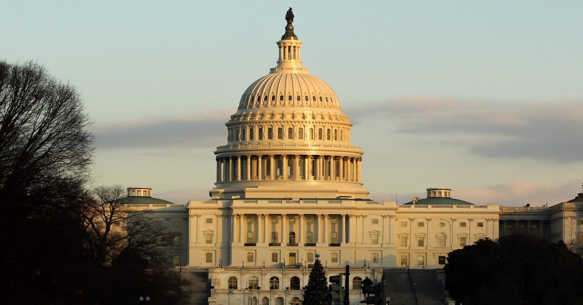 The U.S. Capitol building with its dome illuminated and a Christmas tree in front at sunset.