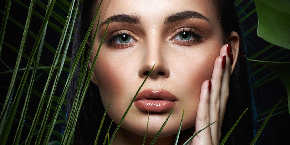 Close-up of a woman with clear skin and glossy lips behind green leaves against a dark background.