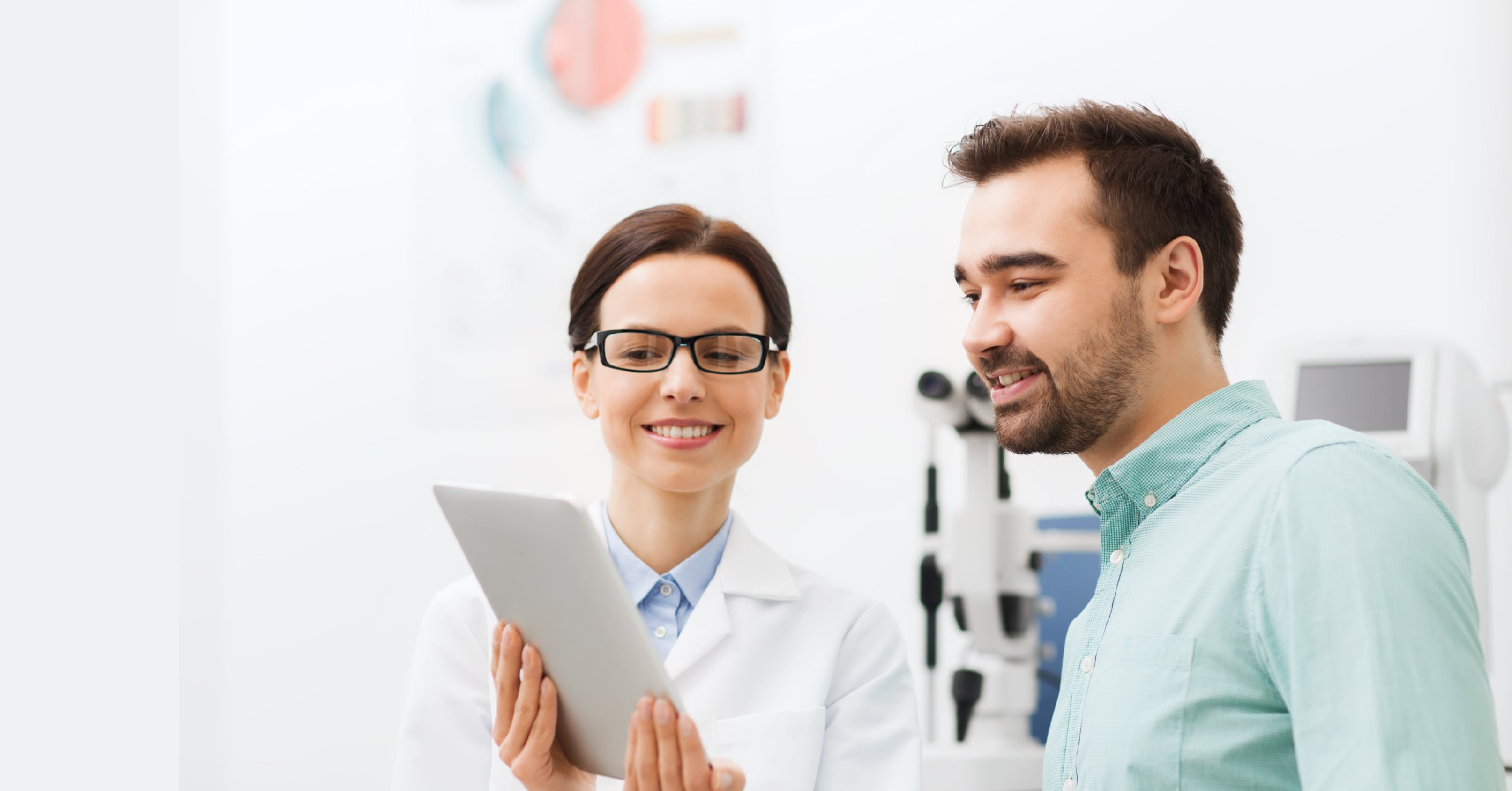 Female eye doctor in white coat with glasses showing a digital tablet to a smiling male patient.