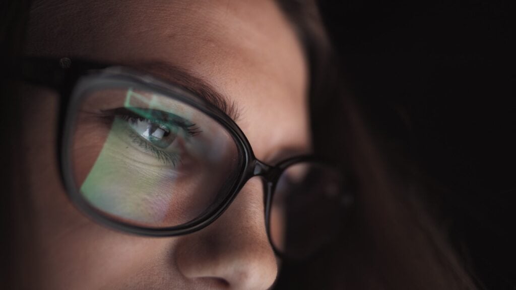 Close-up of a person wearing glasses reflecting a computer screen in a dark environment.