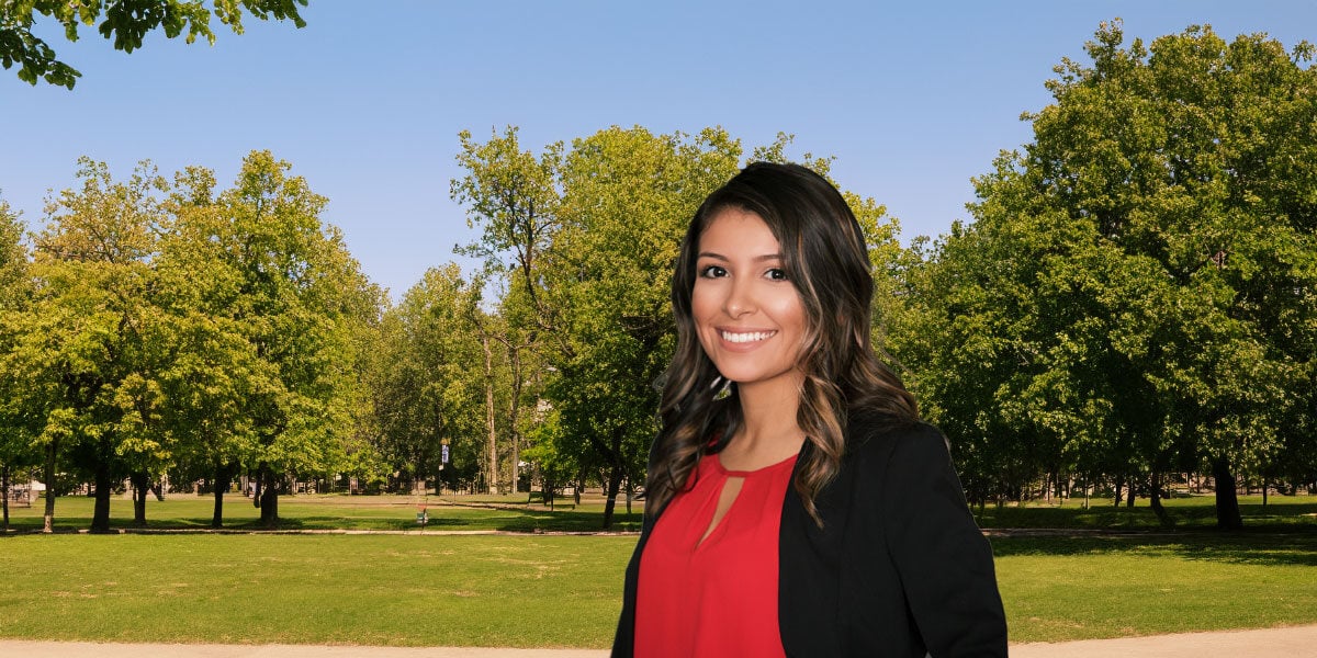 Smiling woman with dark hair wearing a red blouse and black blazer in a green park.