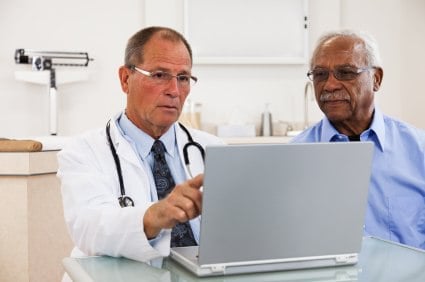 Doctor explaining something on a laptop to an elderly male patient in a clinical setting.