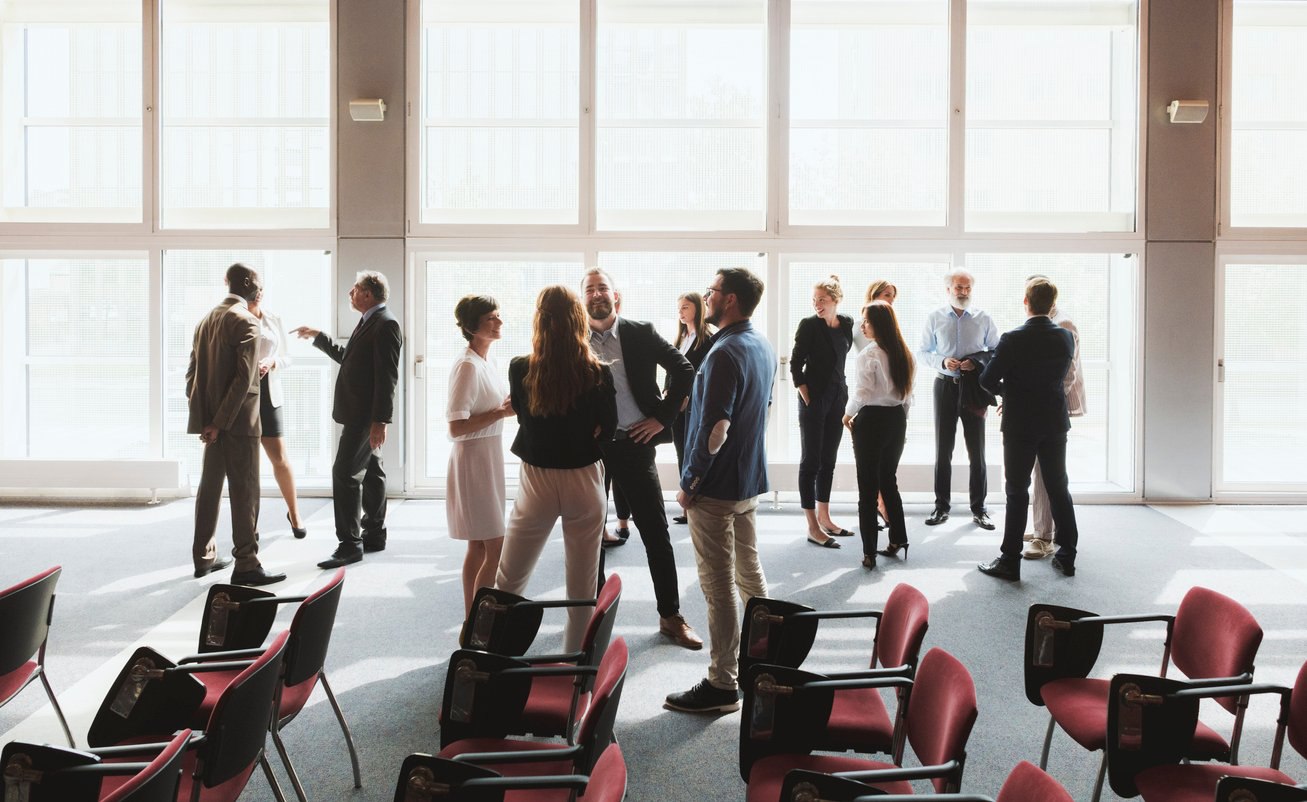 Group of business people standing and talking in a bright conference room with empty chairs.