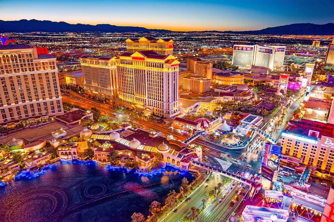 Aerial view of Las Vegas Strip at dusk showing brightly lit casinos, streets, and mountains in the background.