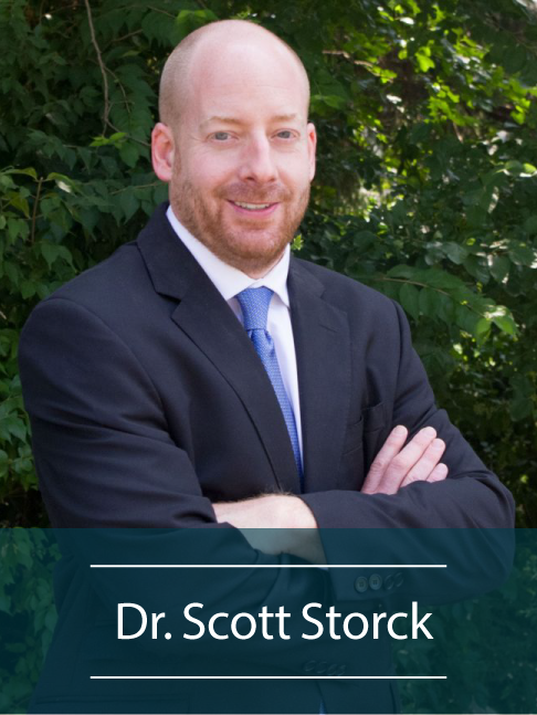Bald man in suit and blue tie standing with arms crossed in front of green leafy background.