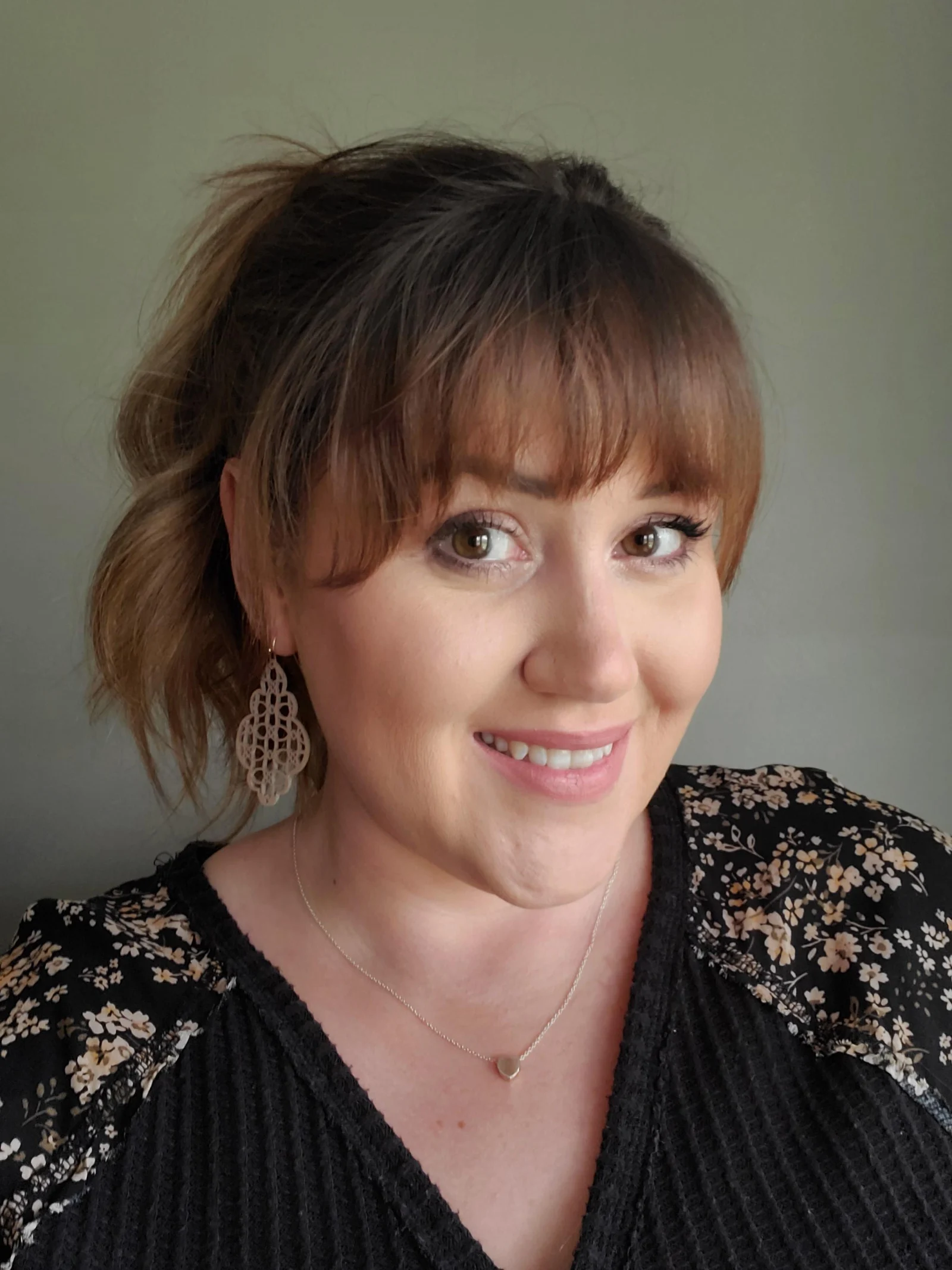 Smiling woman with bangs, wearing floral black top, dangle earrings, and a heart necklace.